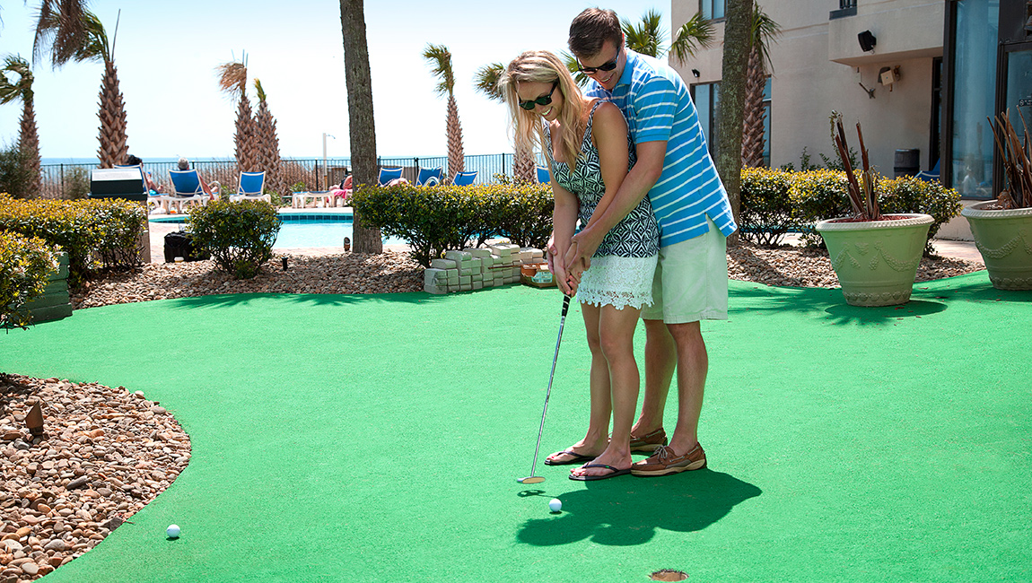 A man and woman play mini golf on a green surface surrounded by palm trees and a pool.